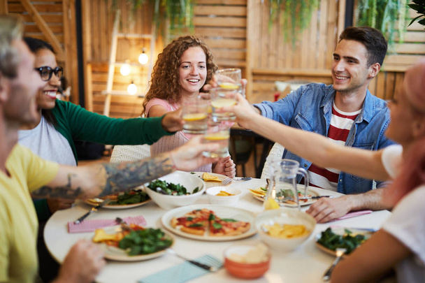 Νέοι ενθουσιασμένοι φίλοι κλίνει με τα ποτά πάνω σερβίρεται γιορτινό τραπέζι, ενώ γιορτάζει εκδήλωση ζωής - Φωτογραφία, εικόνα