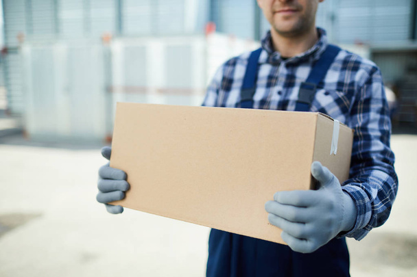 Close-up of moving company worker in gloves holding cardboard box and carrying it at container storage area - Foto, immagini