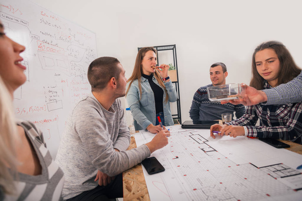 Grupa młodych przedsiębiorców omawiająca swój kolejny projekt na spotkaniu wskazującym plan, który jest na środku stołu. - Zdjęcie, obraz