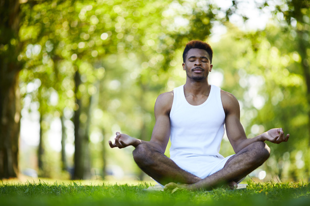 ruhiger, entspannter schwarzer Mann mit Schnurrbart sitzt mit überkreuzten Beinen und hält die Hände in Mudra auf Knien, während er allein im Sommerpark meditiert - Foto, Bild