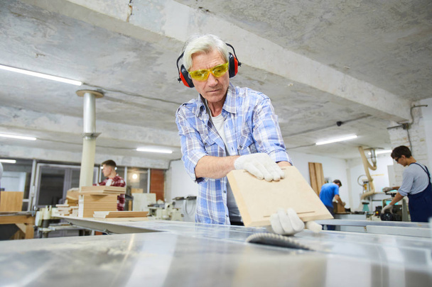 Grave carpintero senior de pelo gris concentrado en guantes de trabajo parado en el escritorio de la sierra de mesa y haciendo tablero de madera en el taller de fábrica
 - Foto, imagen