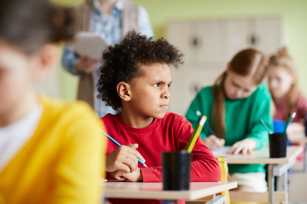 Хмурый задумчивый африканский школьник с вьющимися волосами, сидящий за столом и думающий об испытаниях, глядя в сторону на класс
 - Фото, изображение