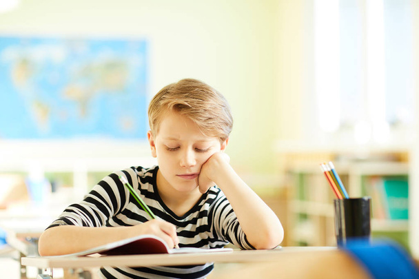 Втомлений незадоволений школяр у роздягненому светрі, що спирається на руку і виконує домашнє завдання або малює в робочій книзі, сидячи за столом у класі
 - Фото, зображення