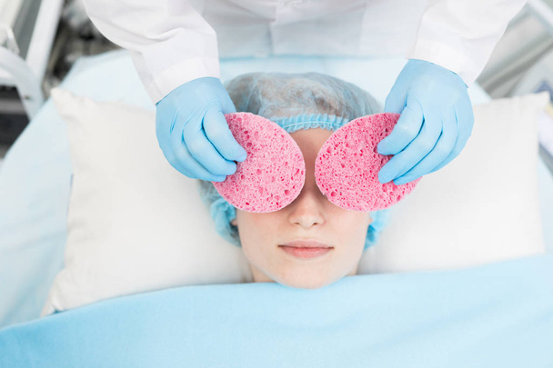 Kosmetolog w niebieskich rękawicach posiadających dwie duże różowe gąbki kosmetyczne na twarz klienta podczas zabiegu pielęgnowania - Zdjęcie, obraz