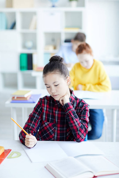 Niña pensativa con lápiz de colores sentada junto al escritorio en el aula y mirando la página en blanco del copybook mientras dibuja
 - Foto, imagen