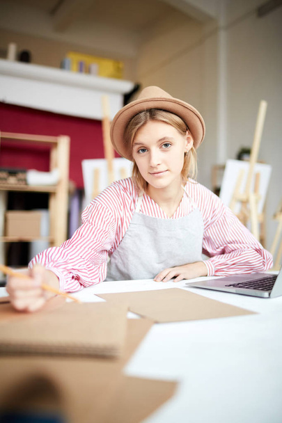 Красивая девушка в шляпе и рабочей одежде сидит на рабочем месте и рисует в блокноте в студии
 - Фото, изображение