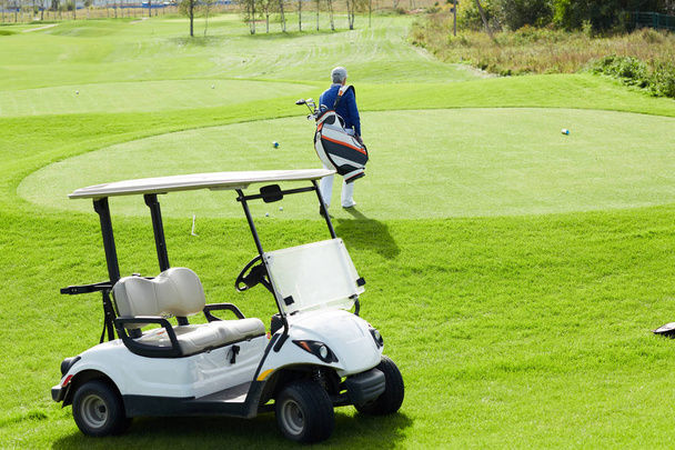Carro de golfe em pé no campo verde e visão traseira do homem idoso com tacos de golfe andando para jogar
 - Foto, Imagem