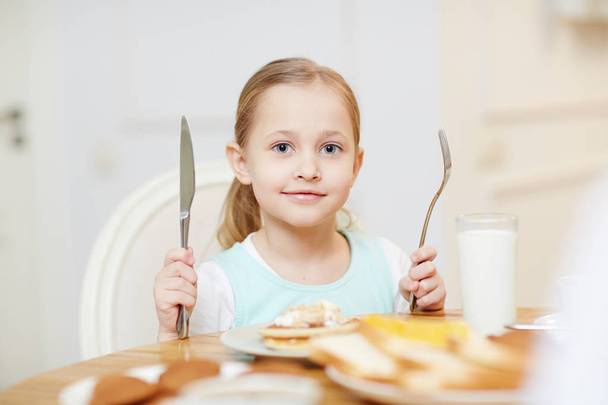 Content pretty kid with blond hair looking at camera and holding fork and knife while ready to eat in kitchen - Foto, Imagen