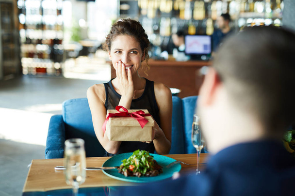 Mladá vzrušená žena držící skříňku s nečekaným dárkem od přítele během oběda v restauraci - Fotografie, Obrázek