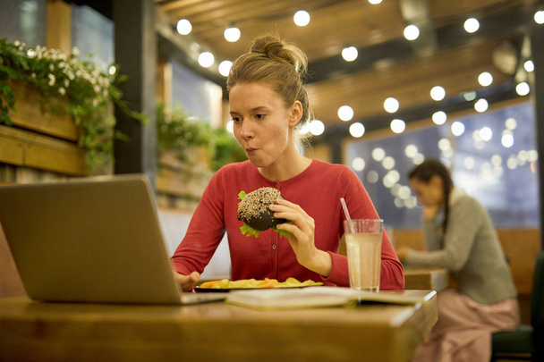 Chica hambrienta comiendo hamburguesa con queso y papas fritas en la cafetería de la universidad mientras busca información en la red
 - Foto, imagen