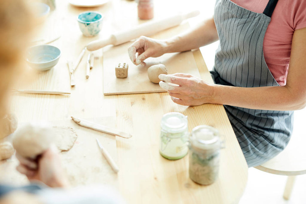 Творческая женщина сидит за деревянным столом с глиняными заготовками и делает кости
 - Фото, изображение