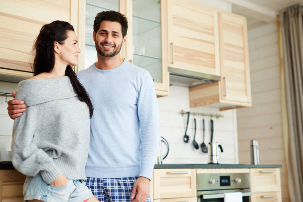 Cheerful handsome young bearded man in pajamas smiling at camera and hugging his girlfriend while they standing in cozy kitchen with wooden set - Foto, imagen