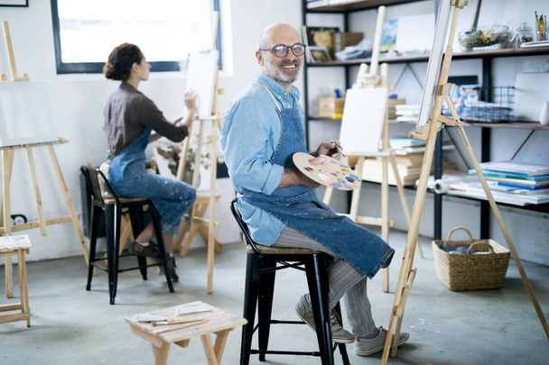 Pintor envelhecido alegre com paleta sentada na frente do cavalete e olhando para você durante o trabalho
 - Foto, Imagem