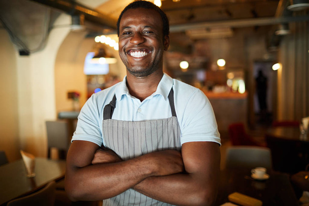 Молодий смішний офіціант з білою зубною посмішкою, що перетинає руки на грудях в кафе
 - Фото, зображення