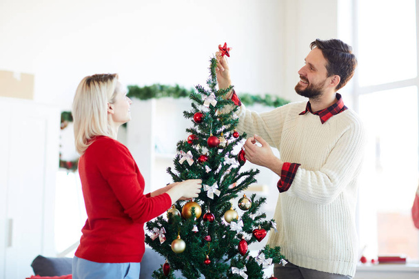 Young man holding decorative red star on top of Christmas tree while talking to his wife - Photo, Image