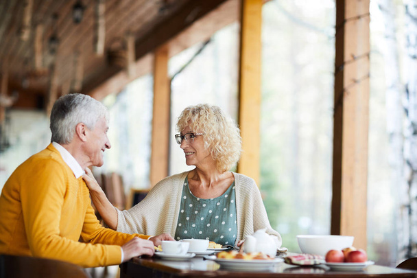 Happy beautiful senior wife with curly hair wearing glasses touching shoulder of husband and supporting him while they talking at breakfast on porch - Photo, Image