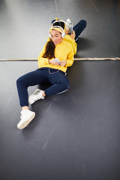 Vista general de la chica descansada en ropa deportiva escuchando música en los auriculares mientras se sienta en el suelo contra un espejo grande
 - Foto, imagen
