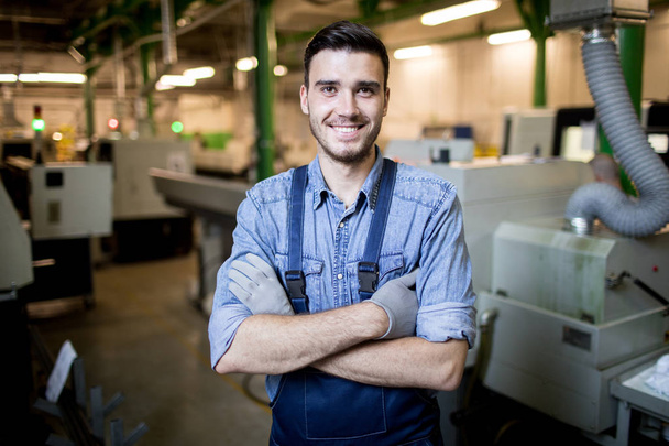 Portrait of young engineer with arms crossed smiling at camera while standing in factory - Valokuva, kuva