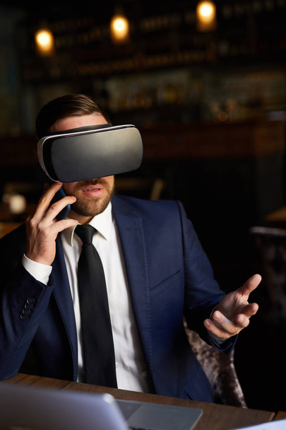 Confident successful bearded businessman watching virtual presentation and discussing it on phone while using virtual reality goggles and gesturing in restaurant - Foto, Imagem