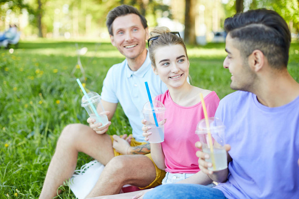 Happy Girl i dwóch facetów z napojami rozmawiając podczas relaksu na piknik w letni dzień - Zdjęcie, obraz