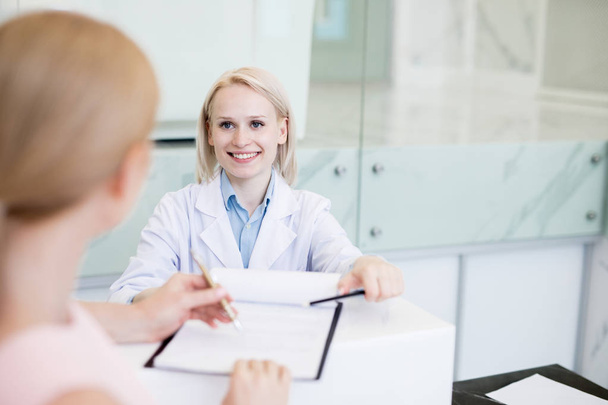 Счастливая молодая женщина в белом халате просит пациента подписать медицинский документ во время консультации
 - Фото, изображение