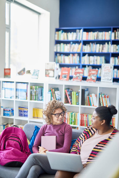 Důvěřiví multietnické studentky sedí na pohovce s brašlemi a diskutují plán projektu v knihovně, seriózní afroameričanka s nadváhou, která během diskuse používá přenosný počítač - Fotografie, Obrázek