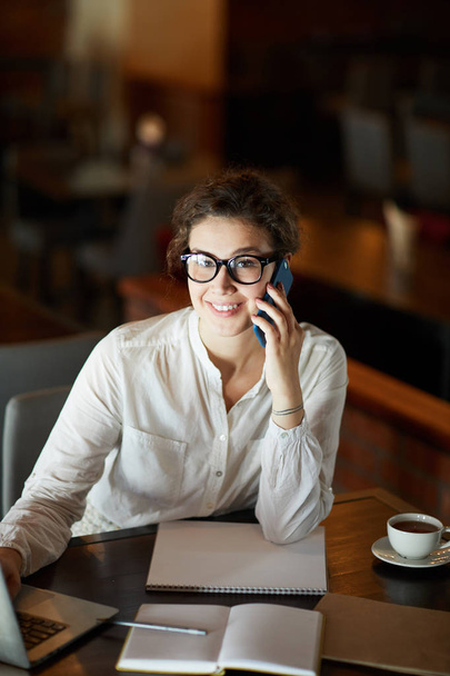 Young smiling businesswoman calling by smartphone while organizing work by table in cafe - Photo, Image