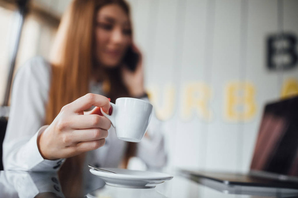 Businesswoman dressed in white blouse with long straight hair sitting at table with cup of coffee, selective focus - Foto, Imagem