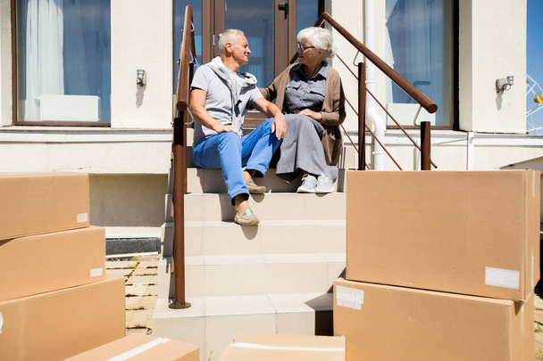 Портрет счастливой пожилой пары, сидящей на крыльце нового дома с картонными коробками на переднем плане, копировальное пространство
 - Фото, изображение