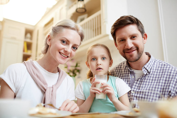 Потрясенная симпатичная дочка с открытым ртом сидит среди родителей и держит полный стакан молока, счастливые члены семьи смотрят на камеру на кухне
 - Фото, изображение