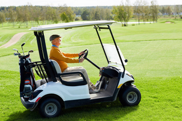 Homem idoso contemporâneo dirigindo carro de golfe ao longo de vasto campo verde enquanto vai para o jogo
 - Foto, Imagem