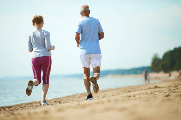 Вид ззаду на старшу пару в активному одязі, що біжить уздовж берегової лінії на піщаному пляжі
 - Фото, зображення