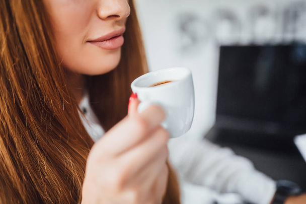 Attractive businesswoman with long straight hair holding cup of coffee near lips and smiling - Фото, зображення