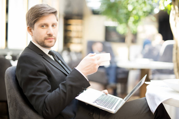Mladý podnikatel s notebookem a šálkem čaje tráví ráno v kavárně tím, že prohlíží online zprávy - Fotografie, Obrázek