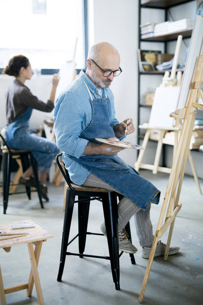 Kypsä mies valitsee värin paletista istuessaan tuolilla maalaustelineen edessä
 - Valokuva, kuva