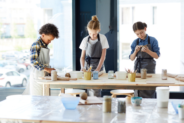 Trois enfants créatifs debout sur le lieu de travail tout en faisant des choses à partir d'argile dans l'atelier
 - Photo, image