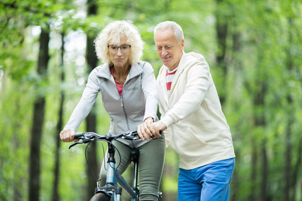 Aged man teaching his wife how to ride bicycle while chilling in natural environment - Photo, Image