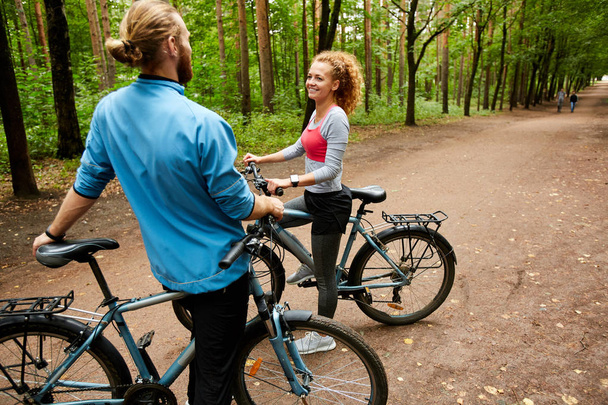 Happy young active couple with bicycles having talk while standing on forest road on summer deay - Photo, Image
