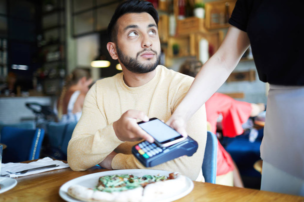 Современный парень сидит за столом в кафе, глядя на официанта, делая бесконтактную оплату через свой смартфон
 - Фото, изображение