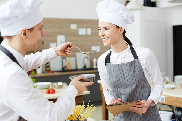 Sorrindo bela jovem mulher em chapéu de chefs e avental degustação de alimentos de interno cozinheiro, jovem dando colher com molho para examinador
 - Foto, Imagem