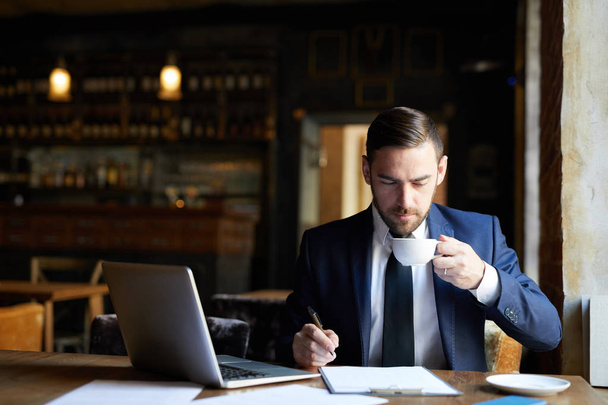 Serious handsome young bearded businessman drinking coffee and examining document while working in fashionable restaurant, he preparing report - Photo, Image