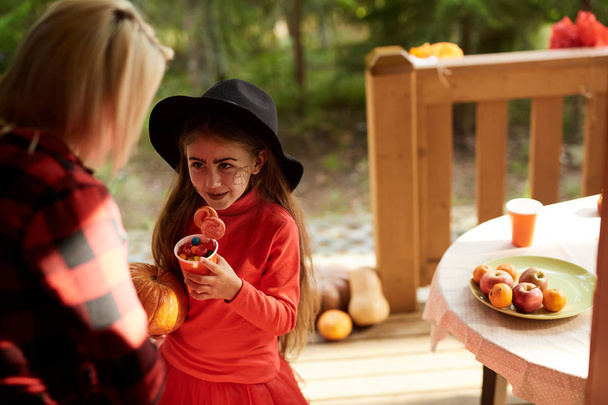Cute halloween girl in hat and red attire holding sweets and talking to young woman - Photo, Image
