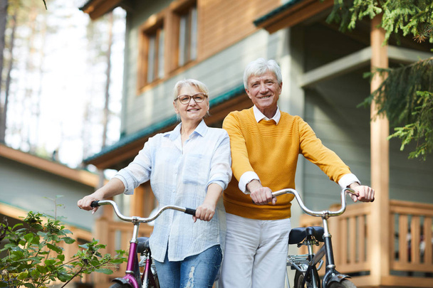 Веселі активні старші пари в повсякденному одязі, що стоять проти заміського будинку і тримають велосипеди, дивлячись на камеру
 - Фото, зображення