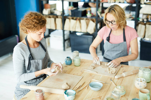 Jonge vrouw en haar collega in schorten zitten bij houten tafel en werken met klei werkstukken - Foto, afbeelding
