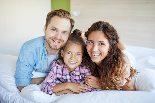 Young laughing restful couple and their daughter in casualwear lying on bed - Fotoğraf, Görsel