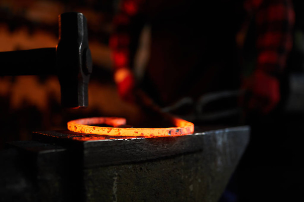 Martillo sobre pieza de trabajo de metal fundido en caliente en yunque con herrero en el fondo
 - Foto, Imagen
