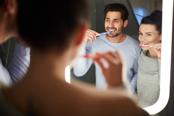 Веселая молодая пара в домашней одежде смотрит в зеркало и веселится, чистя зубы вместе в ванной комнате
 - Фото, изображение
