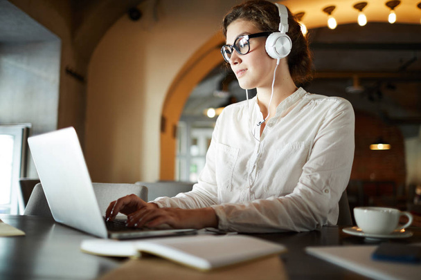 Estudante contemporâneo com fones de ouvido digitando no laptop e ouvindo música enquanto rede no café
 - Foto, Imagem