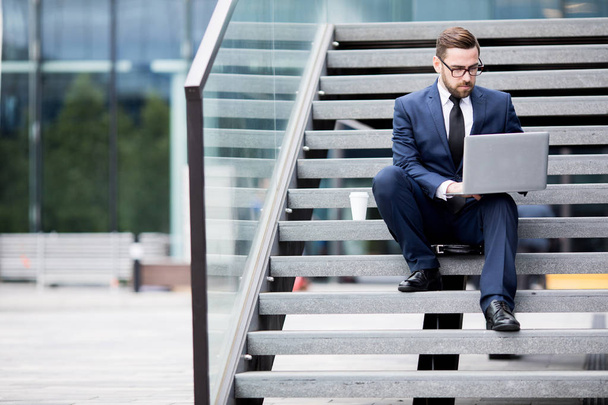 Young stylish male in business dress, eyeglasses looking laptop sitting on rungs  - Foto, immagini