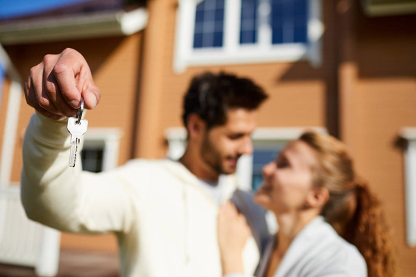 Νέος άνθρωπος δείχνει το κλειδί του νέου σπιτιού, ενώ κοιτάζοντας τη γυναίκα του σε εξωτερικούς χώρους - Φωτογραφία, εικόνα